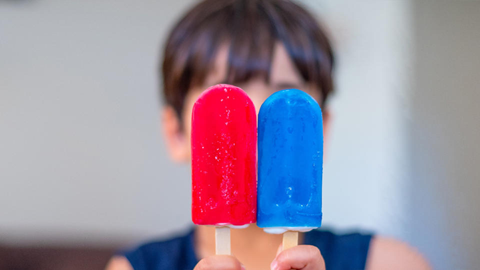 A boy holding red and blue popsicles. 