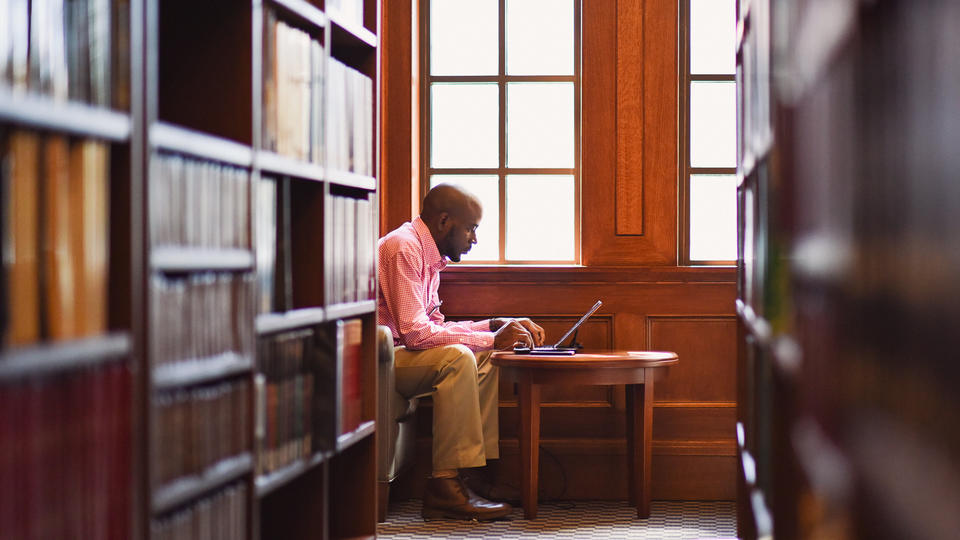 male student in pink button-down and khakis working on computer in the library