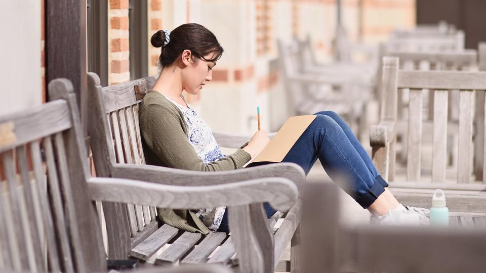 student sitting on benches in courtyard writing in notebook
