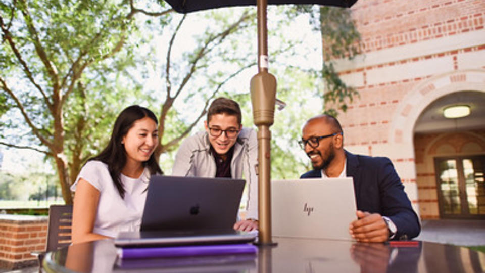 Three students around a small table with umbrella all looking at laptops and smiling