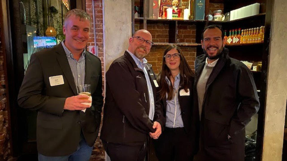 Four alumni standing in a line holding drinks in a dark bar