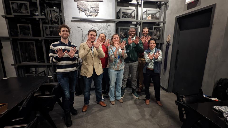 Group of alumni standing in a dimly lit dining room with hands up imitating owls