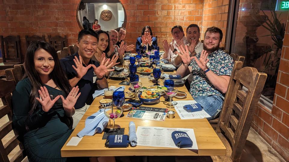 Group of alumni seated around a rectangular table making 'owl hands'