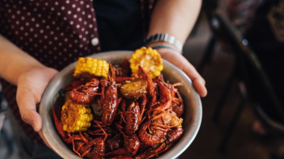 A person in a maroon button-down shirt holding out a bowl of crawfish and corn