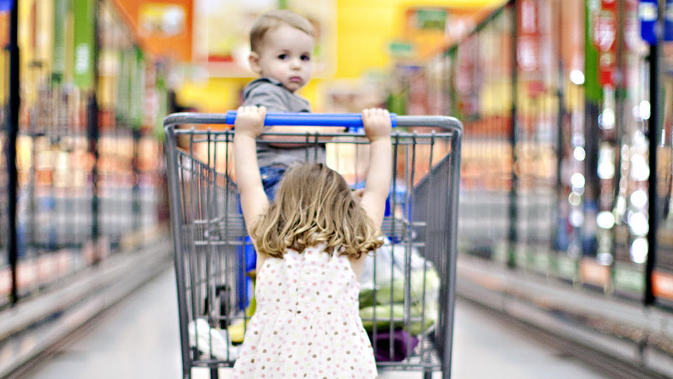 Little girl pushing younger sibling in a grocery cart.
